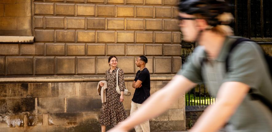 A cyclist wearing a helmet passes a couple talking as they walk in front of a stone wall.