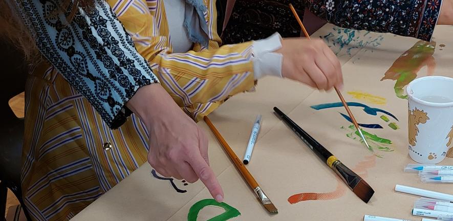 Several hands painting on paper covering a table, with art supplies scattered on top.