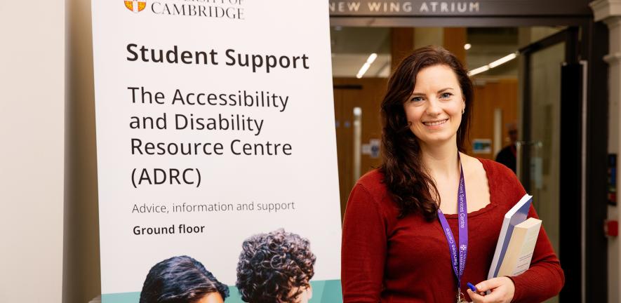A smiling woman holding books and wearing a lanyard stands next to a sign for "Student Support"