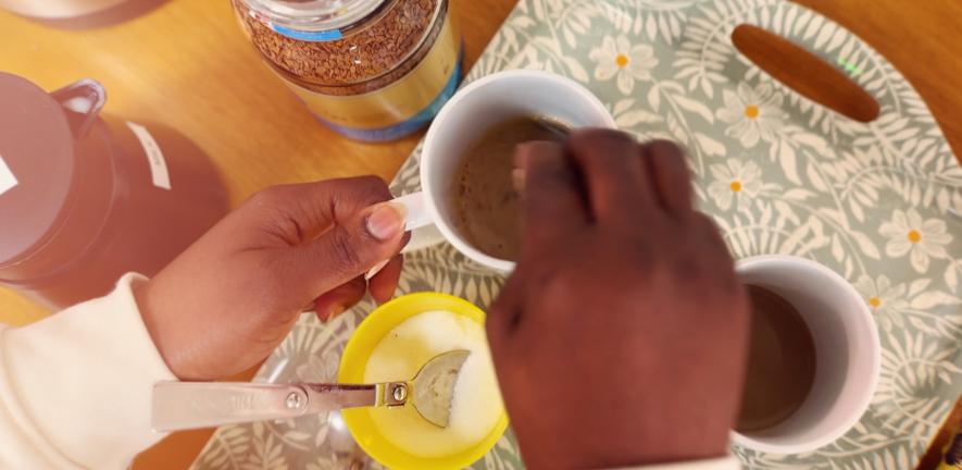 A pair of hands stirring instant coffee in mugs on a flowery tray in sunlight.