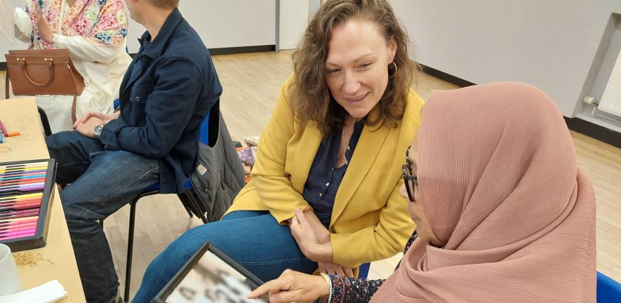 Two women seated at a table covered with art supplies, discussing a family photo, while others talk nearby.