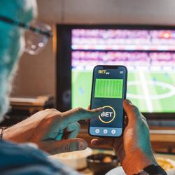 Man sitting on sofa at home and watching a football match. He is using smartphone for sports betting