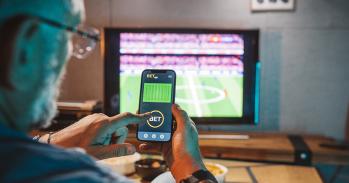 Man sitting on sofa at home and watching a football match. He is using smartphone for sports betting