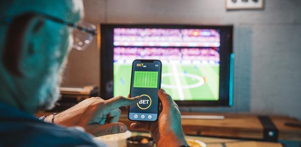 Man watching football and placing a bet, Credit: svetikd via Getty Images Man sitting on sofa at home and watching a football match. He is using smartphone for sports betting