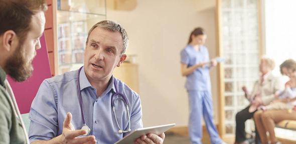 A male doctor sits next to a male patient in a waiting room while holding a digital tablet. In the background a nurse chats to a family while holding digital tablet.