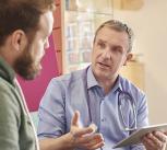 A male doctor sits next to a male patient in a waiting room while holding a digital tablet. In the background a nurse chats to a family while holding digital tablet.