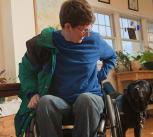Woman with multiple sclerosis in a wheelchair putting on her coat with service dog watching her