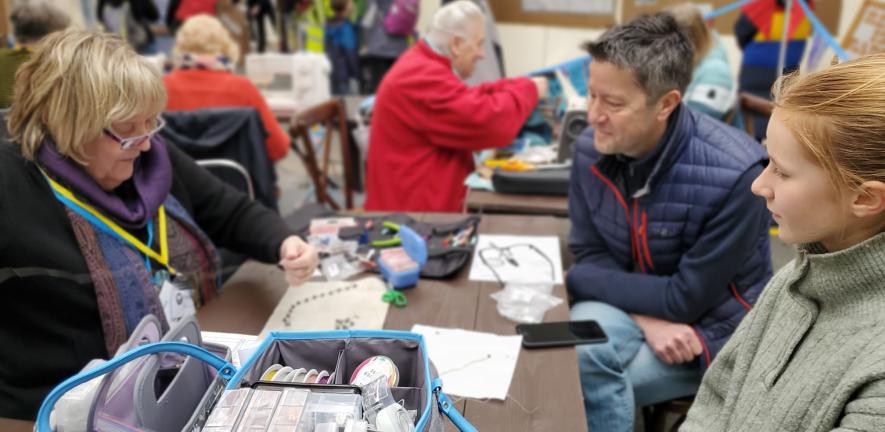 Lots of people interacting at a community event. In the foreground three people are sewing at a table.