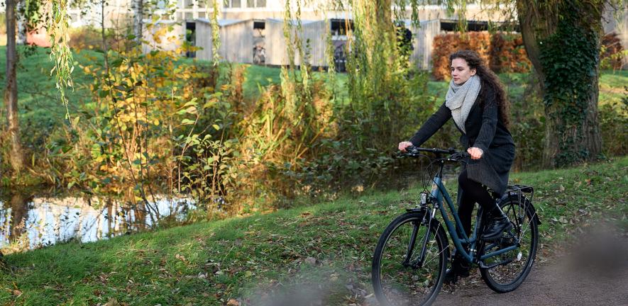 A woman wearing a coat and scarf cycles alongside a small lake