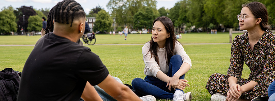 Three students sitting on the grass at Jesus Green in conversation.